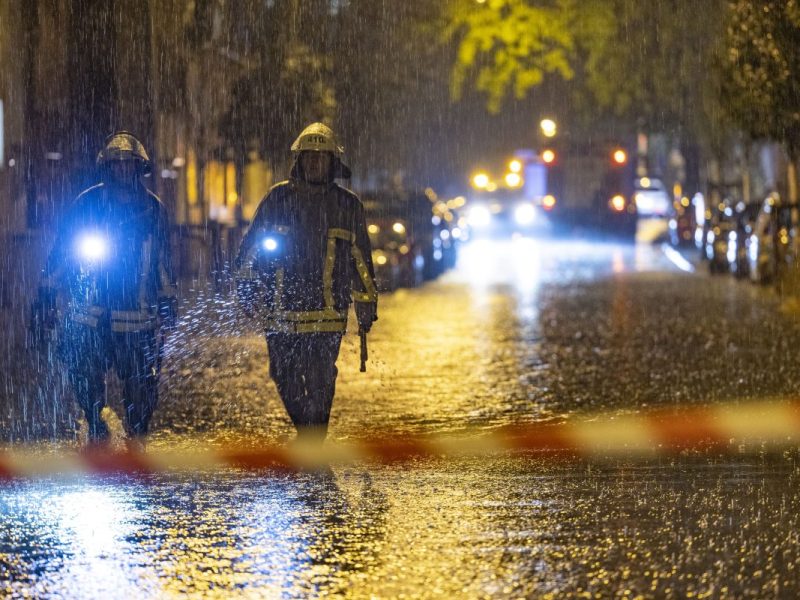 Zwei Feuerwehrmänner laufen in Duisburg über eine überschwemmte Straße mit Flatterband