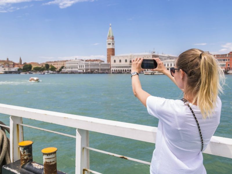 Eine Frau ist auf einem Schiff und steht an der Reling. Sie macht mit ihrem Handy ein Foto von einer Sehenswürdigkeit.