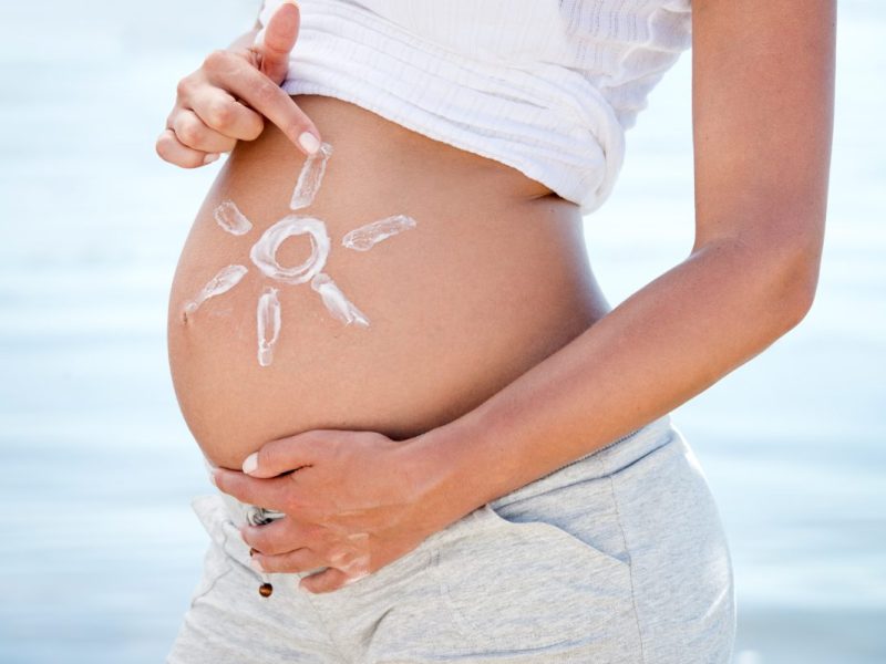 Schwangerschaftsbauch von der Seite. Die Person hält ihre Hände an den Bauch. Mit einer Hand malt sie eine Sonne mit Sonnencreme auf die Seite vom Bauch.