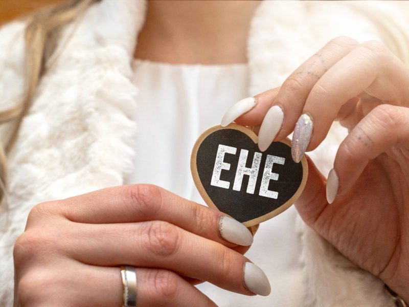 Es ist ein herzförmiges Schild im Vordergrund, auf dem im weißer Schrift "Ehe" steht. Das Schild ist schwarz und hat eine braune Umrandung. Es wird gehalten, von einer Frau mit weißen Nägeln, sie trägt einen Ehering in Silber.