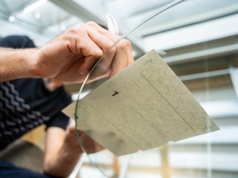 Das Bild wurde durch einen Glastisch von unten Fotografiert. Auf dem Tisch liegt ein Blattpapier, welches mit einem Stift unterschrieben wird. Man sieht eine Hand, welche den Stift hält.